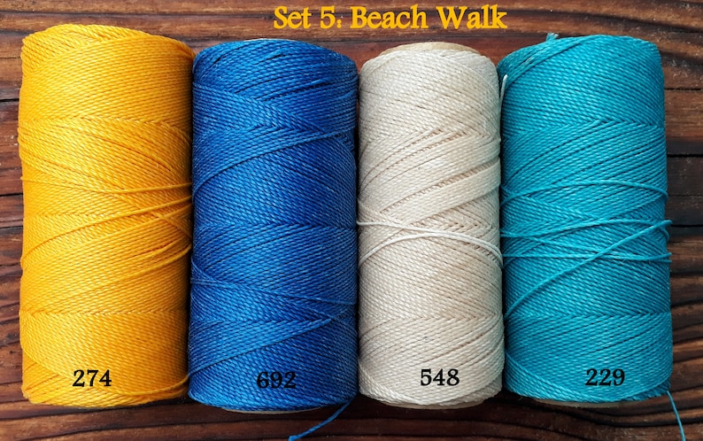 Puede incluir: Conjunto de cuatro carretes de hilo de bordar en amarillo, azul, blanco y turquesa. Los carretes est&aacute;n etiquetados con los n&uacute;meros 274, 692, 548 y 229. El texto "Set 5: Beach Walk" es visible en la parte superior de la imagen.