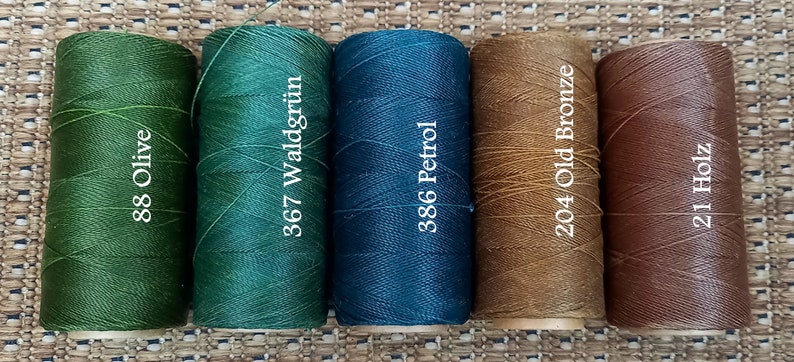 May include: Five spools of waxed thread in different colors. The spools are labeled with numbers and color names: 88 Olive, 367 Waldgr&uuml;n, 386 Petrol, 204 Old Bronze, and 211 Holz.