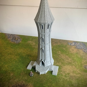 May include: A gray, 3D-printed fantasy tower with a pointed roof and arched windows. The tower is set on a stone base and sits on a green and brown surface. A small figurine is at the base of the tower.