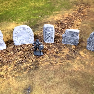 May include: A collection of gray, stone-like monuments with engraved symbols and a miniature warrior figure. The monuments vary in shape and size, with intricate carvings. The scene is set on a brown and green surface, suggesting a tabletop game.