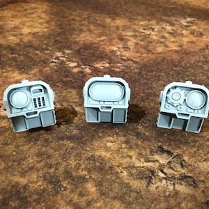 May include: Three light gray miniature components with detailed designs. Each has a rectangular base and a top section with circular and rectangular features. One has a dial, another a screen, and the third a gear.