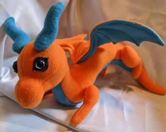 May include: A plush dragon toy featuring bright orange fabric with contrasting light blue horns, wings, and underbelly. The creature has large, dark, glossy eyes and a long, slender body with four limbs, crafted from soft material.
