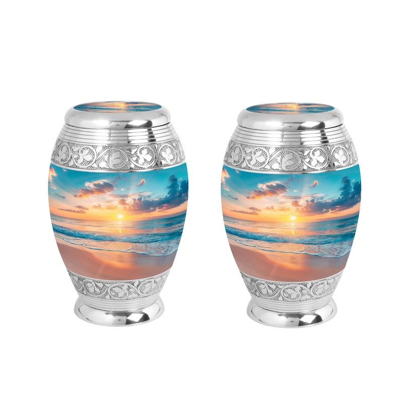 May include: Two silver urns with a beach sunset image. The urns have a decorative floral design around the top and bottom.