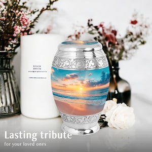 May include: Silver cremation urn with a beach sunset scene. The urn has an ornate silver design around the top and bottom.