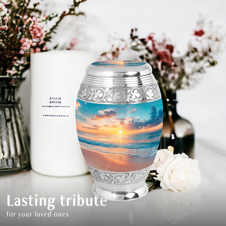 May include: Silver cremation urn with a beach sunset scene. The urn has an ornate silver design around the top and bottom.