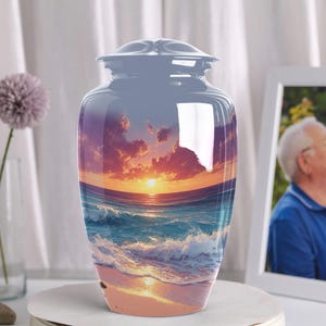 May include: A glossy, blue cremation urn with a sunset and ocean scene. The urn has a lid and a wide, rounded shape. The image on the urn features a vibrant sunset over the ocean with waves crashing on the shore. The urn is displayed on a white surface.