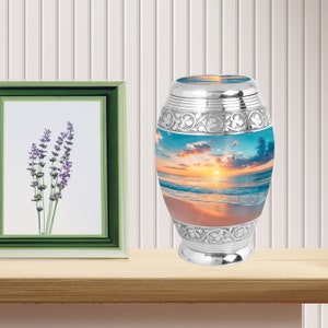 May include: Silver cremation urn with a beach sunset scene. The urn has a silver lid and a detailed floral design around the base.