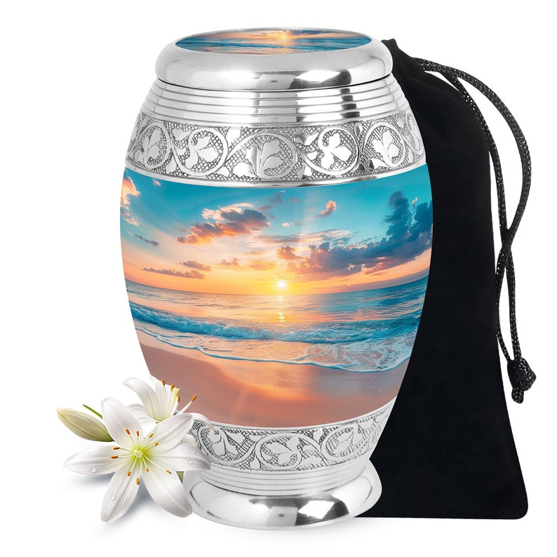 May include: Silver cremation urn with a beach sunset image. The urn has an ornate design and comes with a black velvet bag.