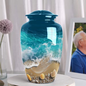 May include: A teal cremation urn with a beach scene design. The urn features a glossy finish with a depiction of ocean waves, sand, and seashells. The urn is displayed with a framed photo and decorative flowers.