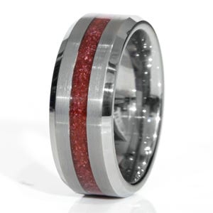 May include: A silver-colored ring with a central band of sparkling red material. The ring has a polished, reflective surface and a brushed metal finish on the outer bands. The design is modern and geometric, suitable for everyday wear.