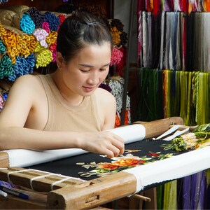 May include: A woman is embroidering a floral design on black fabric using a wooden embroidery hoop. The hoop is set up on a table with a white cloth underneath. The woman is wearing a beige tank top and has her hair pulled back. There are spools of colorful thread in the background.