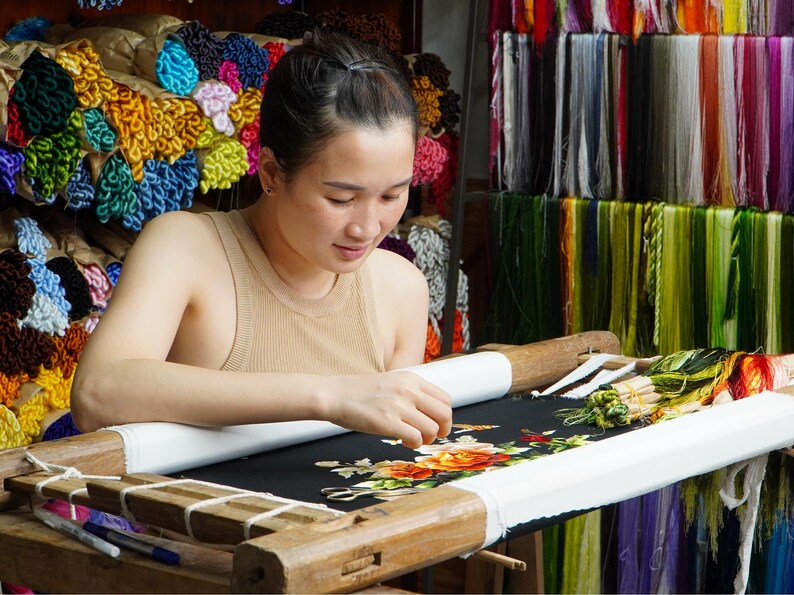 May include: A woman is embroidering a floral design on black fabric using a wooden embroidery hoop. The hoop is set up on a table with a white cloth underneath. The woman is wearing a beige tank top and has her hair pulled back. There are spools of colorful thread in the background.