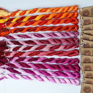 May include: A collection of 18 skeins of hand-dyed silk embroidery floss in shades of orange, red, pink, and white. The skeins are arranged in a gradient from orange to white. Each skein has a brown tag with the text "THUONG LONG EMBROIDERY" printed on it.