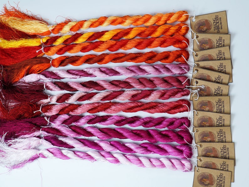 May include: A collection of 18 skeins of hand-dyed silk embroidery floss in shades of orange, red, pink, and white. The skeins are arranged in a gradient from orange to white. Each skein has a brown tag with the text "THUONG LONG EMBROIDERY" printed on it.