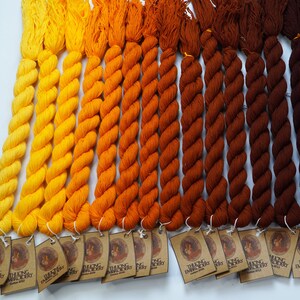 May include: A collection of 15 skeins of embroidery floss in shades of yellow, orange, brown, and blue. The floss is arranged in a gradient from light yellow to dark blue. Each skein has a label with the text "THUONG EMBROIDERY" and a small image of a woman.