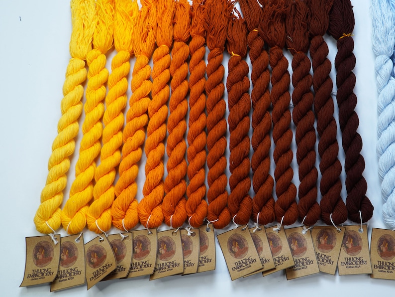 May include: A collection of 15 skeins of embroidery floss in shades of yellow, orange, brown, and blue. The floss is arranged in a gradient from light yellow to dark blue. Each skein has a label with the text "THUONG EMBROIDERY" and a small image of a woman.