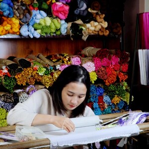 May include: A woman sits at a table working on an embroidery project. She is using a variety of colorful threads to create a design. The table is covered in a white cloth and there are several spools of thread on the table. The background is filled with shelves of colorful thread.
