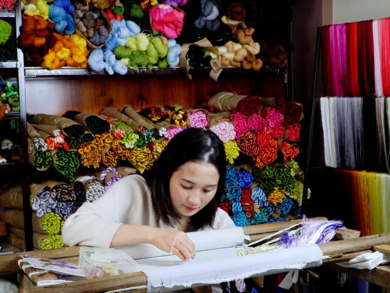 May include: A woman sits at a table working on an embroidery project. She is using a variety of colorful threads to create a design. The table is covered in a white cloth and there are several spools of thread on the table. The background is filled with shelves of colorful thread.