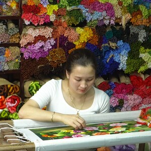 May include: A woman sits at a table, working on a floral embroidery project. She is surrounded by spools of colorful thread, creating a vibrant backdrop. The embroidery features a detailed design of roses and leaves.