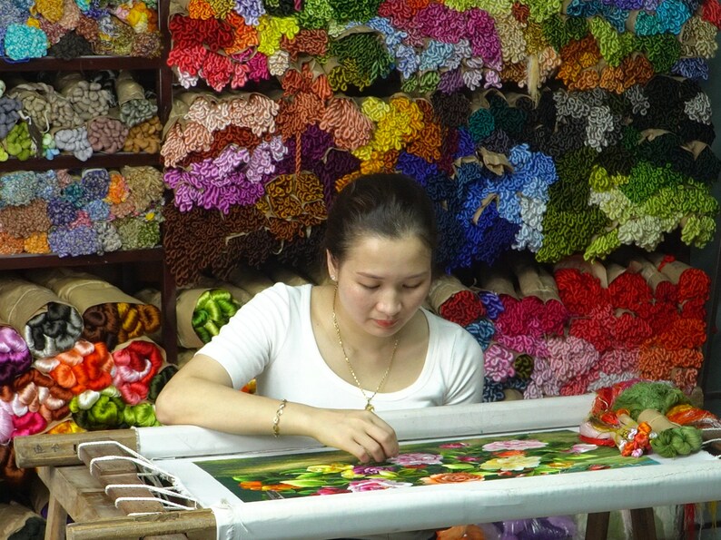 May include: A woman sits at a table, working on a floral embroidery project. She is surrounded by spools of colorful thread, creating a vibrant backdrop. The embroidery features a detailed design of roses and leaves.