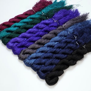May include: A collection of six skeins of hand-dyed silk thread in various shades of blue, green, and purple. The skeins are arranged in a fan shape, with the darkest shade at the bottom and the lightest shade at the top.