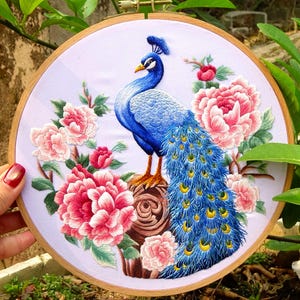 Puede incluir: Arte de bordado en aro con un pavo real azul con una larga cola de plumas iridiscentes. El pavo real está de pie en una rama con flores rosas.