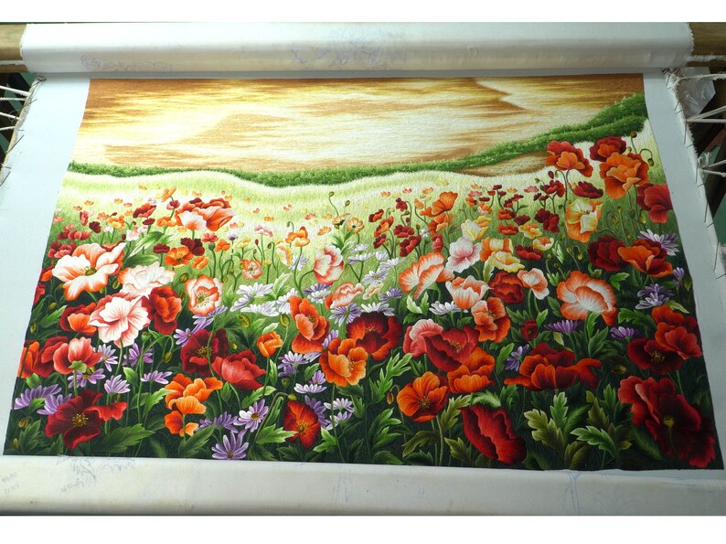 May include: A close-up of a needlepoint embroidery depicting a field of red, orange, and white poppies with green leaves and stems. The background is a soft yellow and brown, suggesting a sunset or sunrise.