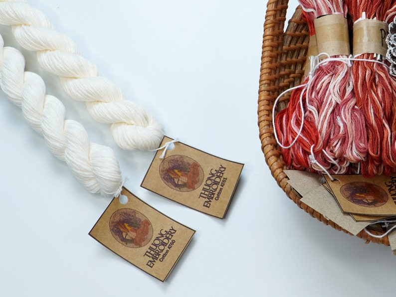 May include: Two spools of white cotton embroidery floss with brown paper tags that read "THUONG EMBROIDERY Cotton 100%". A wicker basket holds several spools of red and white embroidery floss.