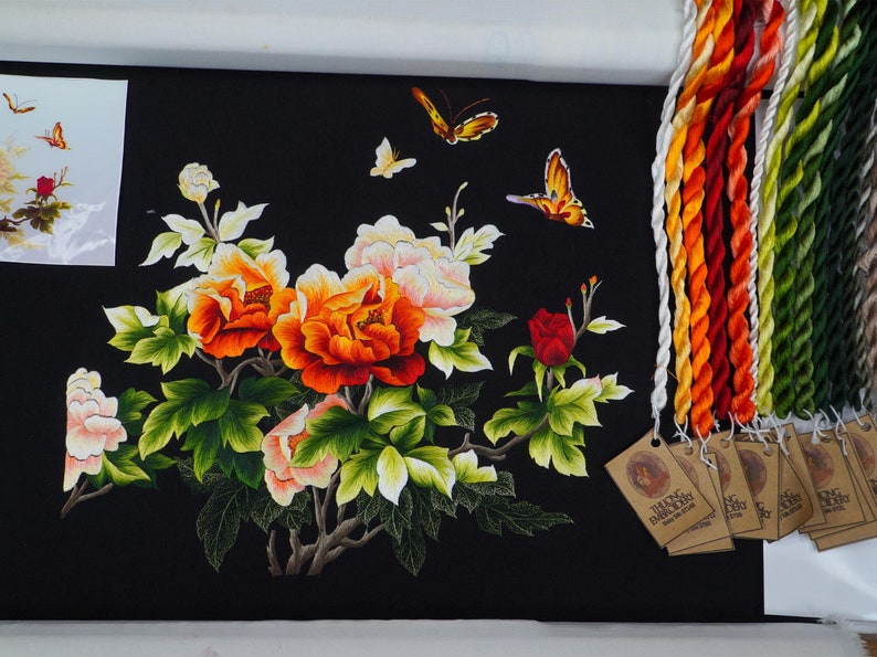 May include: Embroidered artwork featuring a floral design with red, orange, and white peonies, green leaves, and butterflies on a black background. The artwork is framed and includes a set of embroidery floss.