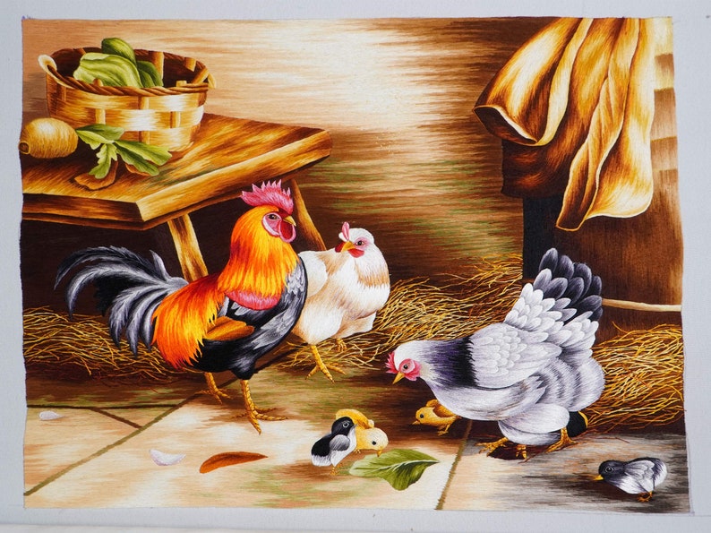 May include: A colorful embroidered painting depicting a rooster, two hens, and three chicks. The rooster is black, orange, and yellow with a red comb. The hens are white and gray. The chicks are yellow and black. The painting is set against a brown background with a wooden table and a brown cloth.