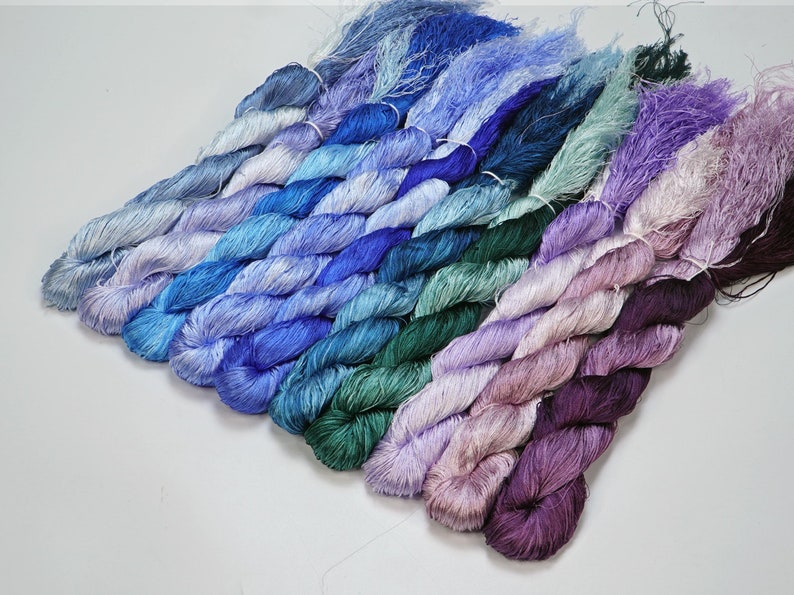 May include: A collection of 10 skeins of hand-dyed silk thread in shades of blue, green, purple, and pink. The skeins are arranged in a rainbow pattern.