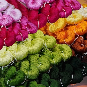 May include: A variety of colorful yarn spools stacked in a rainbow pattern. The yarn is arranged in rows of different colors, including pink, red, yellow, orange, brown, green, and dark green.