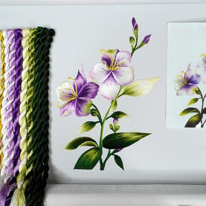 May include: A close-up of a needlepoint embroidery project featuring a white and purple flower with green leaves. The embroidery is on a white fabric and is surrounded by a wooden frame. There are several spools of embroidery floss in various colors of purple, green, and white.