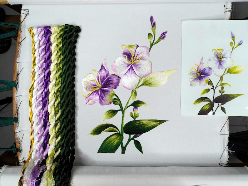 May include: A close-up of a needlepoint embroidery project featuring a white and purple flower with green leaves. The embroidery is on a white fabric and is surrounded by a wooden frame. There are several spools of embroidery floss in various colors of purple, green, and white.
