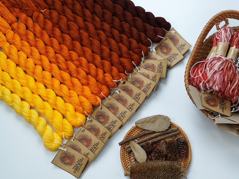 May include: A collection of 12 skeins of hand-dyed yarn in shades of orange, yellow, and brown. The yarn is arranged in a gradient from dark brown to light yellow. Each skein has a small brown tag with the text "The Indigo Dragon" printed on it.
