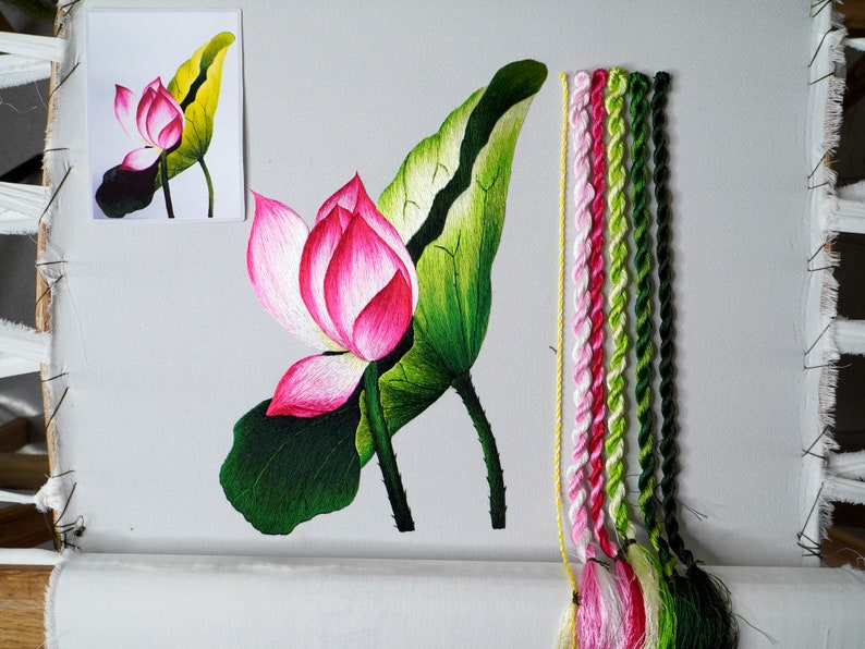 May include: Embroidered lotus flower with green leaves and a pink blossom. The embroidery is done on white fabric and the floss is in shades of pink, green, and yellow.