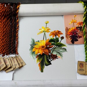 May include: A close-up of a needlepoint embroidery project featuring a bouquet of yellow and orange flowers with green leaves. The embroidery is on a white canvas and is surrounded by a wooden frame. There are several skeins of embroidery floss in various shades of orange, brown, and green.