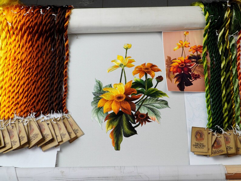 May include: A close-up of a needlepoint embroidery project featuring a bouquet of yellow and orange flowers with green leaves. The embroidery is on a white canvas and is surrounded by a wooden frame. There are several skeins of embroidery floss in various shades of orange, brown, and green.