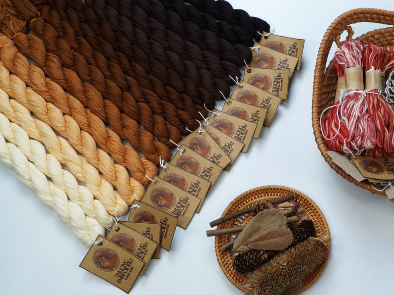 May include: A collection of 16 skeins of embroidery floss in shades of brown, orange, and cream. The floss is arranged in a gradient from dark brown to cream. Each skein has a brown paper tag with the text "Talking Embroidery" and "Cotton Embroidery Floss".