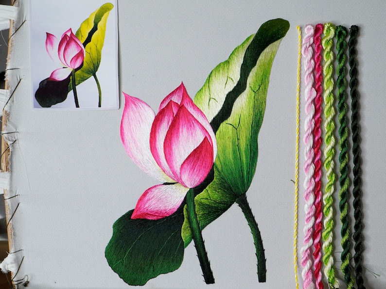 May include: Embroidery project featuring a pink and white lotus flower with green leaves. The embroidery is done on white fabric and uses a variety of colored threads.