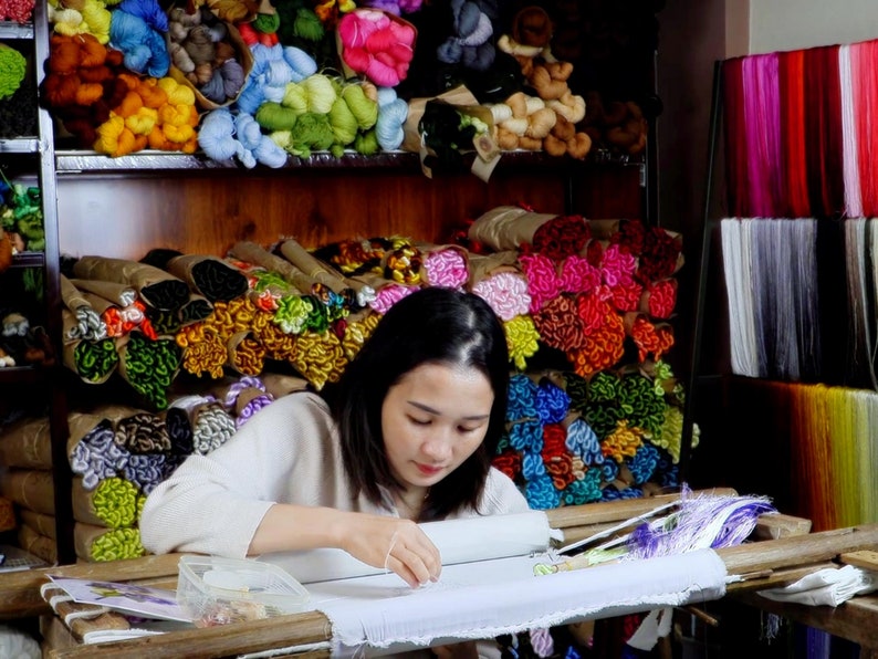 May include: A woman sits at a table working on an embroidery project. She is using a variety of colorful threads to create a design. The table is covered in a white cloth and there are several spools of thread on the table. The background is filled with shelves of colorful thread.