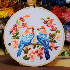 Peut inclure: Art de broderie en forme de cercle représentant deux oiseaux bleus avec des becs orange perchés sur une branche avec des fleurs roses et vertes. Les oiseaux sont entourés d'un fond blanc.