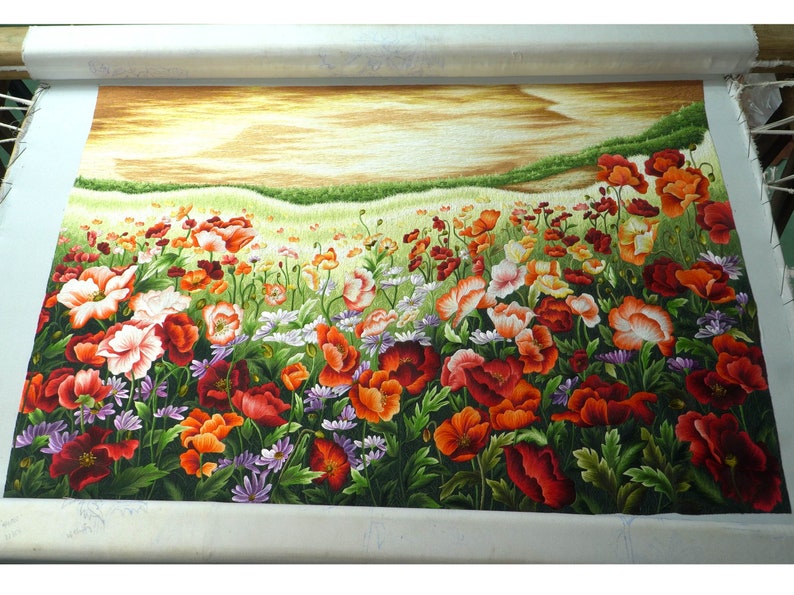May include: A close-up of a needlepoint embroidery depicting a field of red, orange, and white poppies with green leaves and stems. The background is a soft yellow and brown, suggesting a sunset or sunrise.