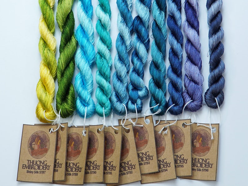 May include: A collection of 10 skeins of embroidery floss in shades of yellow, green, blue, and purple. Each skein is labeled with the brand name "Thuong Embroidery" and a unique product code.