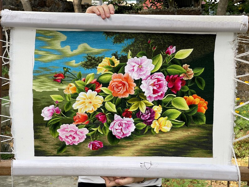 May include: A framed embroidery depicting a bouquet of colorful flowers, including pink, red, yellow, and orange roses, with green leaves and a blue sky background.