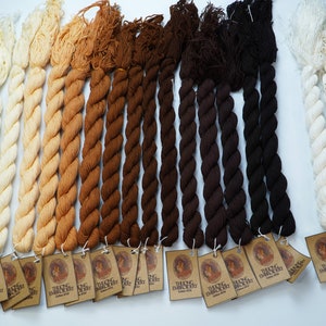 May include: A collection of 10 skeins of cotton embroidery floss in various shades of brown, beige, and white. The floss is arranged in rows, with the darkest shades at the bottom and the lightest shades at the top. Each skein has a small tag attached to it with the words "Thaing Embroidery Cotton 40/2" printed on it.
