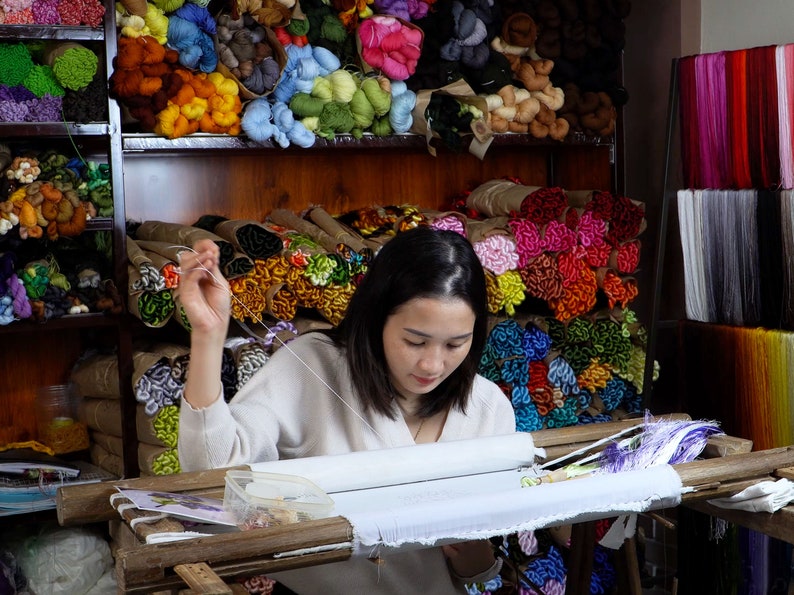 May include: A woman sits at a table with a wooden frame and a white canvas stretched across it. She is using a needle and thread to embroider a design on the canvas. The table is surrounded by shelves filled with colorful spools of thread.