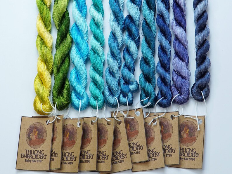 May include: A collection of 10 skeins of embroidery floss in shades of yellow, green, blue, and purple. Each skein is labeled with the brand name "Thuong Embroidery" and a unique product code.
