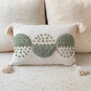 Puede incluir: Una almohada blanca con un diseño geométrico verde. La almohada tiene un ribete de flecos y borlas.