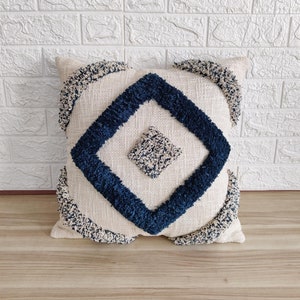 May include: A cream coloured throw pillow with a navy blue diamond pattern and fringe trim. The pillow has a tufted texture and a geometric design.